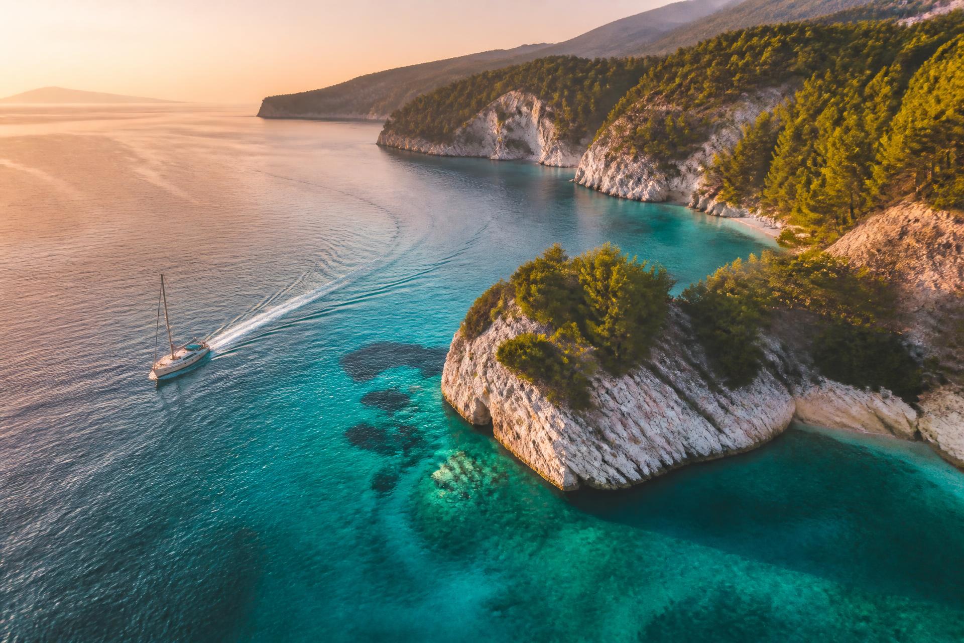 Sailboat crossing turquoise water along a pine-covered coastline at sunset